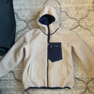Beige teddyjacka från Patagonia - Säljer en mysig beige teddyjacka från Patagonia med mörkblå detaljer. Jackan har en praktisk dragkedja och en bröstficka med dragkedja. Perfekt för kyliga dagar med sin varma och sköna känsla.