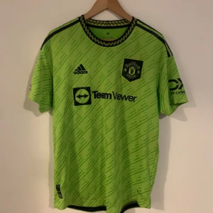 Grön Manchester United  fotbollströja från Adidas authentic - Säljer en grön fotbollströja från Adidas med svarta detaljer och tryck. Tröjan har korta ärmar och ett mönster över hela tyget. Perfekt för fotbollsfans! Nr 18 Casemiro storlek L