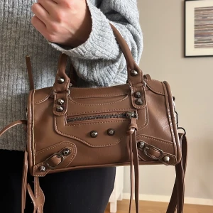 Brun axelväska med dragkedja - Snygg brun axelväska i skinn med  detaljer och dragkedja framtill. Väskan har en praktisk storlek och justerbar axelrem.