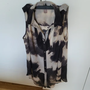 Mönstrad ärmlös blus - Säljer en unik ärmlös blus med ett abstrakt mönster i svart och beige. Blusen har en klassisk krage och knappar framtill, vilket ger en stilren look. Perfekt för en avslappnad men ändå chic stil.