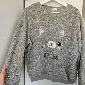 Grå fleece tröja med kattmotiv - Mysig grå fleece tröja med ett gulligt kattmotiv och texten 'Let's Cuddle'. Perfekt för kalla dagar när du vill hålla dig varm och bekväm. Tröjan har långa ärmar och en rund halsringning.