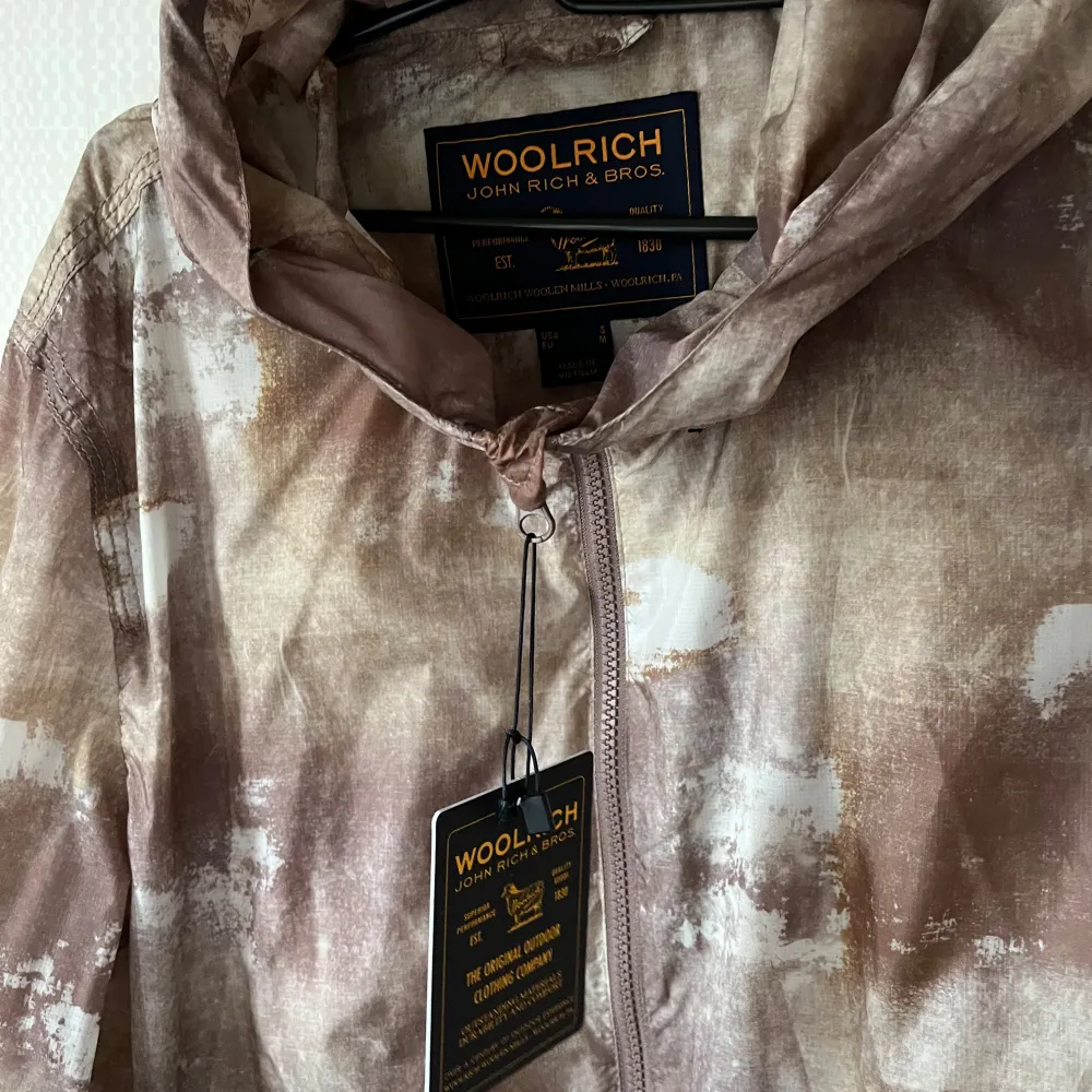 Säljer en cool mönstrad jacka från Woolrich i jordnära toner. Jackan har en dragkedja framtill och en praktisk huva. Perfekt för en avslappnad stil.. Takit.