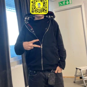 Svart hoodie med dragkedja - Säljer en svart hoodie med dragkedja och rutigt mönster i luvan. Perfekt för en avslappnad stil. Den har långa ärmar och en bekväm passform.