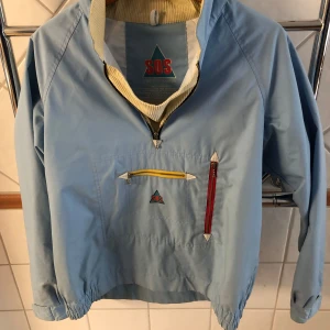 Ljusblå vindjacka från SOS - Retro ljusblå vindjacka från SOS med halv dragkedja framtill och två fickor med färgglada dragkedjor. Jackan har resår i ärmslut och nederkant samt en hög krage med vit och gul insida. Perfekt för dig som gillar vintage stil.