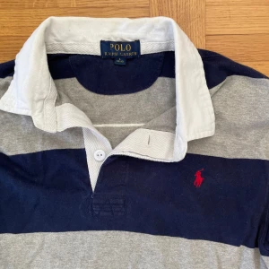 Randig pikétröja från Polo Ralph Lauren - Säljer en långärmad pikétröja från Polo Ralph Lauren i storlek L 14-16 men passar xs och S. Tröjan har breda marinblå och grå ränder, vit krage och en röd broderad logga på bröstet. Stor vit siffra 3 på ryggen. Perfekt för våren och sommaren.