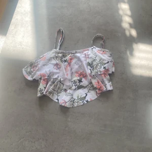 Blommig bikini överdel  - Säljer en söt blommig bikini överdel  med volanger. Överdelen har justerbara axelband och ett vackert blommönster i rosa och grönt på en vit bakgrund. Perfekt för sommardagar!