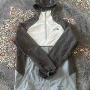Grå och vit vindjacka från The North Face - Säljer en stilren grå och vit vindjacka från The North Face. Jackan har en huva och en halv dragkedja framtill. Perfekt för blåsiga dagar och har en sportig look.
