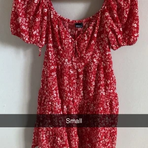Röd blommig klänning med puffärmar - Charmig röd klänning med blommigt mönster och puffiga kortärmar. Den har en söt knytning framtill och en luftig passform som passar perfekt för varma dagar. Perfekt för en somrig look! 🌸
