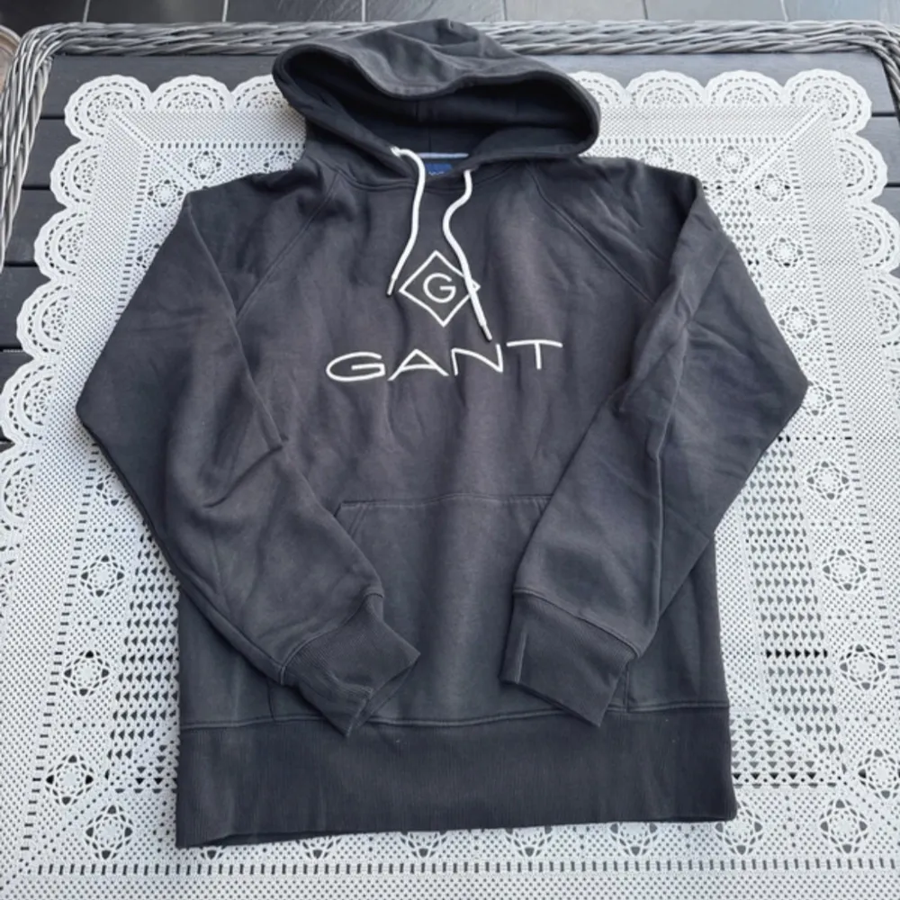 Säljer en stilren svart hoodie från GANT med vit logga på bröstet. Tröjan har en klassisk känguruficka och justerbar huva med vita snören. Perfekt för en avslappnad look.. Neuletakit & Villapaidat.