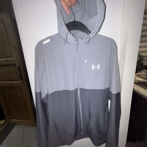 Windbreaker från Under Armour - Säljer en grå windbreaker från Under Armour med två nyanser av grått och vit logga på bröstet. Jackan har huva med dragsko och hel dragkedja framtill. Perfekt för träning eller chill.