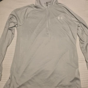 Under Armour halvzip - Silverfärgad långärmad tröja från Under Armour med halv dragkedja och logotyp på bröstet. Perfekt för träning eller vardagsbruk. Tröjan har en lös passform och är tillverkad i ett mjukt material.