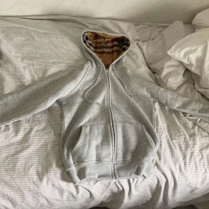 Grå burberry hoodie  - Säljer en grå hoodie som liknar Burberry med dragkedja och klassiskt rutmönstrat foder i huvan. Tröjan har två fickor framtill och ribbade muddar. Perfekt för dig som gillar stilrena och exklusiva plagg. (A-cop) hör av dig vid frågor😊