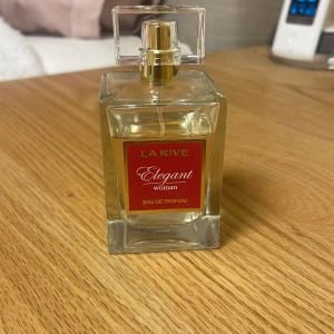 La Rive Elegant Woman Eau de Parfum - En stilren parfym för kvinnor från La Rive, Elegant Woman Eau de Parfum. Flaskan är rektangulär i genomskinligt glas med en guldig spray och ett fyrkantigt lock. Etiketten är röd med guldtext.