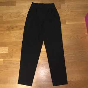 Svarta kostymbyxor - Snygga svarta kostymbyxor med hög midja och raka ben. Perfekta för en stilren look. Byxorna har hällor för bälte och diskreta bakfickor. De är några år gamla men har inte använts mycket, är i bra skick. Saknar dock lappar. Måtten är: midjeomkrets = 68cm, höftomkrets = 96cm, innerbenslängd = 63cm. 