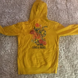 Gul hoodie med tryck från Love & Order - Säljer en gul hoodie från Love & Order med blommigt tryck och orm på ryggen samt liten logga framtill. Tröjan har huva och långa ärmar, perfekt för en färgglad och avslappnad stil.