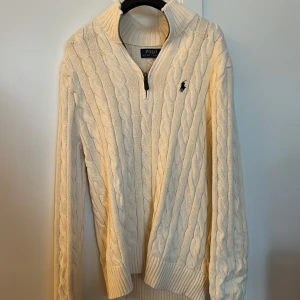 Vit kabelstickad tröja från Polo Ralph Lauren - Kabelstickad vit tröja från Polo Ralph Lauren med halv dragkedja och hög krage. Tröjan har lång ärm, ribbade muddar och den klassiska broderade loggan på bröstet. Perfekt för en stilren och klassisk look.