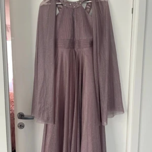 Ljuslila glittrig långklänning med cape och pärldetaljer - Säljer en ljuslila långklänning med glittrigt tyg, cape och pärldetaljer vid halsringningen. Klänningen har plisserad överdel, bred midja och axelbandslös look med cape som ger extra volym. Perfekt för bal eller fest.