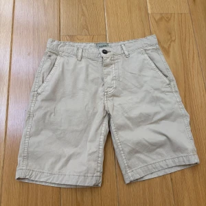 Beiga shorts från Scalpers - Snygga beiga shorts från Scalpers med klassisk passform och broderad döskalle-logga vid fickan. Shortsen har knapp och dragkedja i gylfen samt två sidofickor. Perfekta för varma dagar.