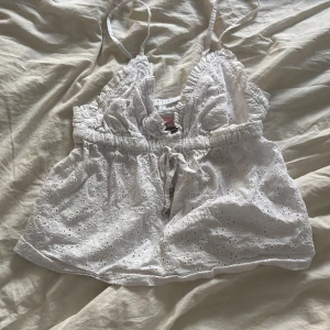 Vit broderad linnetopp från Censored - Söt vit linnetopp från Censored i storlek XS. Toppen har tunna axelband, broderade blommor och en liten rosett framtill. Perfekt för varma dagar och ger en romantisk känsla med sitt luftiga material och fina spetsdetaljer.