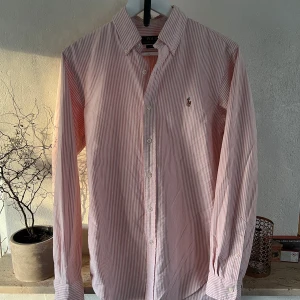 Rosa och vit randig skjorta från Polo Ralph Lauren - Snygg rosa och vit randig skjorta från Polo Ralph Lauren med klassisk krage och broderad logga på bröstet. Skjortan har långa ärmar och knappar framtill. Perfekt för en stilren look.