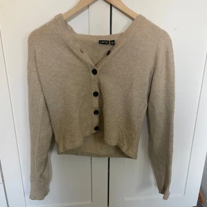 Beige stickad kofta från LMTD - Säljer en beige, mysig stickad kofta från LMTD med svarta knappar framtill. Koftan är lite croppad och har långa ärmar, perfekt för en stilren look. Passar bra till både jeans och kjol. 