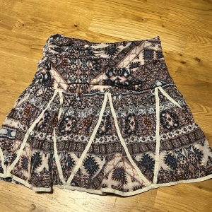 Mönstrad kjol från Bershka - Kort kjol från Bershka med bohemiskt mönster i brunt, blått och beige. Kjolen har dragkedja i sidan och dekorativa sömmar framtill. Lätt och luftig modell, perfekt för varmare dagar.