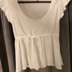 Vit peplumtop med knyt - Söt vit topp med peplumskärning och knyt i midjan. Toppen har korta ärmar och rund halsringning. Den har smink fläck runt halsen men som jag kommer tvätta innan den skickas, toppen är ifrån Nelly i storlek XS knappt använd!!