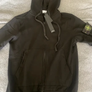 Svart hoodie från Stone Island - Säljer en svart hoodie från Stone Island med dragkedja och huva. Klassisk logga på ärmen och stora fickor framtill. Perfekt för en stilren och avslappnad look. Materialet är mjukt och bekvämt.