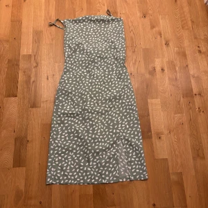 Grön mönstrad långklänning med smala axelband - Säljer en grön långklänning med vita abstrakta prickar och smala axelband som knyts. Klänningen har en rak passform och slits framtill för extra rörelsefrihet. Perfekt för varma dagar och somriga tillfällen.