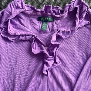 Lila volangblus från Ralph Lauren - Supersöt lila blus från Ralph Lauren med volangdetaljer längs kragen och knäppning framtill. Tillverkad i mjuk bomull, perfekt för dig som vill ha något unikt och färgglatt i garderoben.