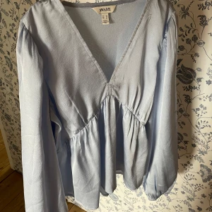 Ljusblå blus med v-ringning från Aware - Säljer en ljusblå blus från Aware med djup v-ringning och långa ärmar. Älskar denna blus och den skimrar så fint i solen men den kommer tyvärr inte till användning! Har en super liten fläck på vänsterarm!!🩵
