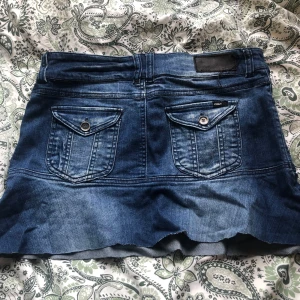 Jeanskjol från ONLY - Säljer en snygg jeanskjol från ONLY i klassisk blå denim. Kjolen har två fickor med lock och knapp bak samt en rå, fransig nederkant. Perfekt för dig som gillar en avslappnad och trendig stil.