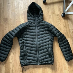  Performance jacka - Säljer en svart dunjacka från Peak Performance i storlek M. Jackan har huva, dragkedja framtill och är quiltad med horisontella sömmar. Perfekt för kyliga dagar och enkel att bära med sig tack vare lättviktsdesignen.