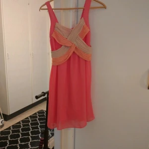 Rosa kortklänning med beige detaljer - Säljer en söt rosa kortklänning med breda axelband och dekorativa beige band framtill. Klänningen har en luftig passform och är perfekt för sommarens tillfällen. Materialet känns lätt och faller fint.