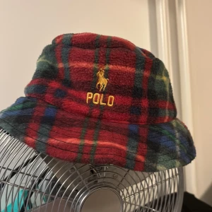 Skön hatt från Polo Ralph Lauren - Snygg och färgglad rutig fleecehatt från Polo Ralph Lauren. Hatten har ett broderat Polo-märke i gult framtill och är perfekt för att hålla värmen med stil. Den har en klassisk bucket hat-design och är tillverkad i mjukt fleece-material.