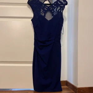 Marinblå klänning med spetsdetaljer - Elegant marinblå klänning med vackra spetsdetaljer vid halsringningen och axlarna. Klänningen har en figurnära passform och en diskret dragkedja i ryggen. 