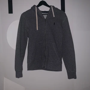 Grå hoodie  från Polo Ralph Lauren - Säljer en grå hoodie från Polo Ralph Lauren med dragkedja och huva. Tröjan har långa ärmar, två fickor framtill och vita snören i huvan. Klassisk liten logga broderad på bröstet.