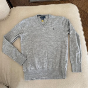 Grå tröja från Polo Ralph Lauren - Grå tunn stickad tröja från Polo Ralph Lauren. Tröjan har en v-ringad design och är tillverkad i ett mjukt material. Perfekt för en klassisk och avslappnad look. Storlek XL/TG 18-20 - small. 