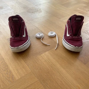 Vinröda sneakers från Vans - Säljer ett par vinröda Vans sneakers med vit sula och klassisk snörning. Skorna har en ikonisk design med vit sidodetalj och är perfekta för en avslappnad stil. Sulan har det typiska rutmönstret för bra grepp.