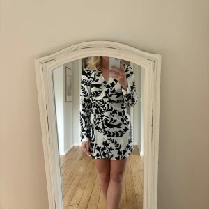 klänning - Snygg kort klänning med svartvitt mönster och knytband i midjan. Klänningen har långa ärmar och en v-ringad hals. Perfekt för en stilren look.