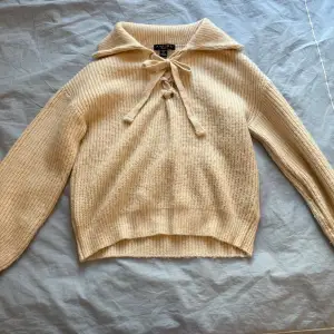 Säljer en beige stickad tröja från Amisu i storlek XS. Tröjan har lång ärm, krage och snygg knytning framtill med metallöljetter. Materialet är en mjuk blandning av akryl och återvunnen polyester. Perfekt för kyla sommarkvällar med en kjol eller över en klänning, eller till vardags med jeans! 