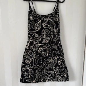 Svart blommig kortklänning med smala axelband - Säljer en svart kortklänning med vita blommiga mönster och smala axelband. Klänningen har en figurnära passform och är perfekt för varma dagar. Enkel och stilren design som passar till många tillfällen.
