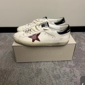 Vita sneakers med röd stjärna från Golden Goose - Säljer ett par vita sneakers från Golden Goose med röd stjärndetalj på sidan och svarta detaljer vid hälen. Skorna har snörning och rund tå. Perfekta för dig som gillar en avslappnad och trendig stil.