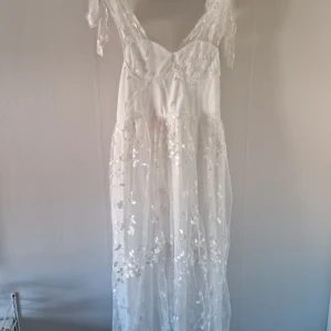 Vit långklänning med spets och blommigt mönster - Romantisk vit långklänning med tunna axelband och vacker spetsdetalj. Klänningen har ett transparent lager med blommigt mönster och volang vid axelbanden. Perfekt för sommarens speciella tillfällen.