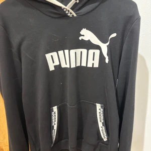 Svart hoodie från Puma - Svart hoodie från Puma med stor vit logga och text på bröstet. Tröjan har huva med svartvitt band och en stor magficka framtill med Puma-detaljer. Perfekt för en sportig och avslappnad stil.
