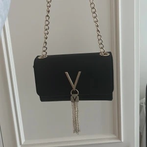 Svart axelväska från Valentino - Säljer en svart axelväska från Valentino med guldfärgad kedja och stor V-detalj framtill. Väskan har dekorativa kedjor som hänger ner och stängs med magnetlås. Perfekt för att lyfta din outfit med en lyxig touch.