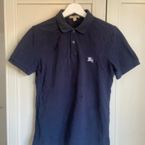 Mörkblå pikétröja från Burberry - Säljer en klassisk mörkblå pikétröja från Burberry i 100% bomull. Tröjan har korta ärmar, krage och knäppning framtill samt diskret Burberry-logga broderad på bröstet. Perfekt för en stilren och avslappnad look.