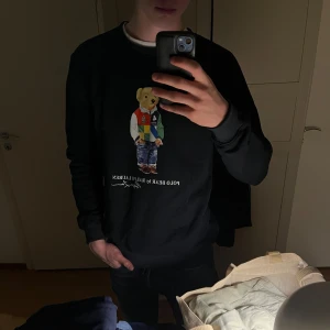 Svart sweatshirt med Polo Bear från Ralph Lauren - Svart sweatshirt från Ralph Lauren med den klassiska Polo Bear-figuren på bröstet. Tröjan har rund halsringning och långa ärmar. Perfekt för en avslappnad och stilren look.