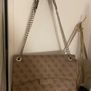 Beige axelväska från Guess med kedjerem - Säljer en beige axelväska från Guess med logomönster över hela väskan. Väskan har en lång kedjerem i silverfärg och lock med magnetknäppning. Perfekt för att bära dina viktigaste saker med stil. Lite sliten i bandet 