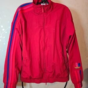 Röd Adidas vindjacka med blå och rosa ränder - Säljer en röd vindjacka från Adidas med dragkedja och klassiska blå och rosa ränder längs ärmarna. Jackan har broderad Adidas-logga på ena ärmen och två fickor framtill. Perfekt för en sportig look.